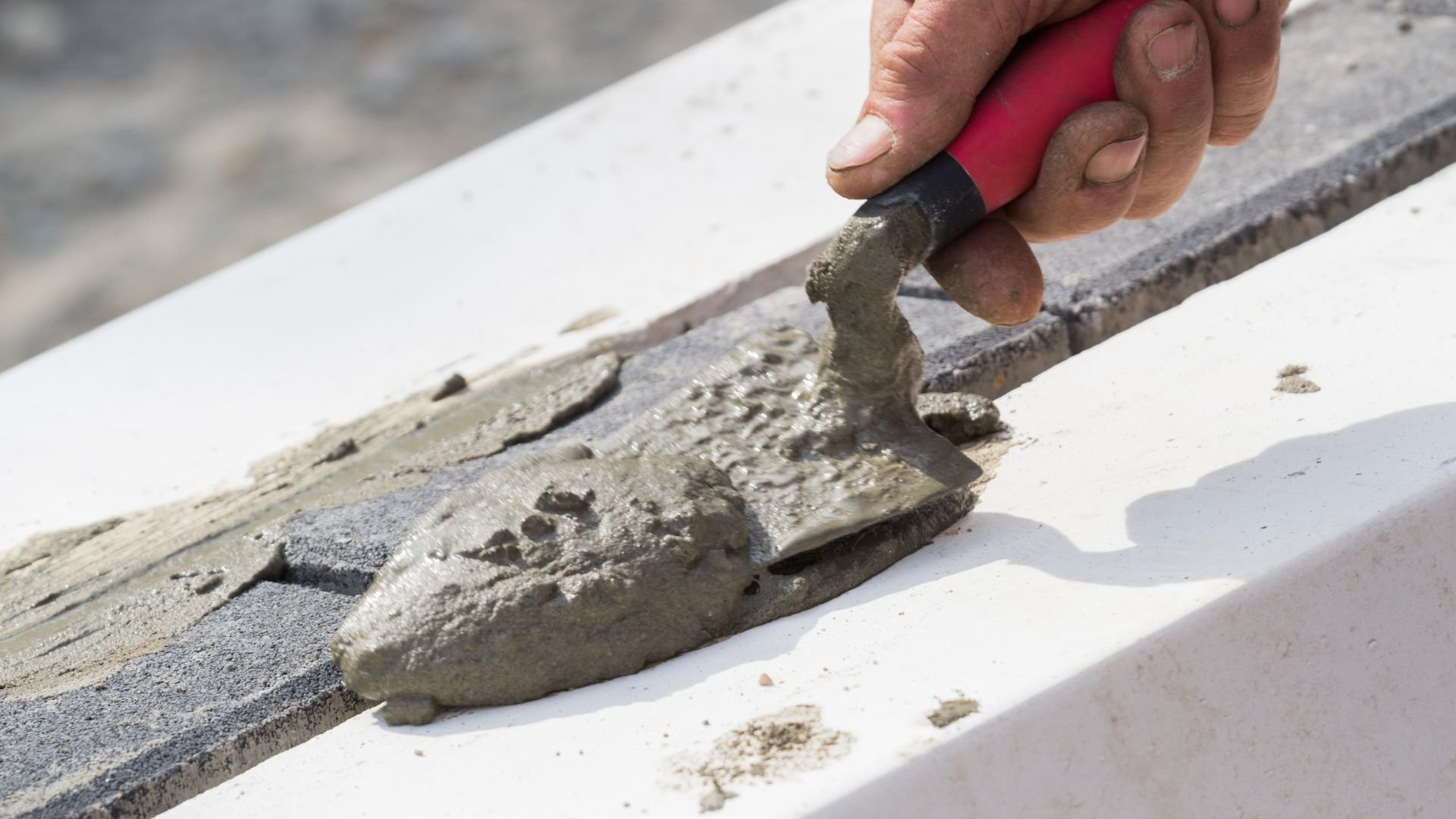 Hand applying wet cement with red-handled trowel on construction surface
